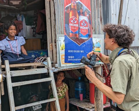 VuTheara dans un bidonville avec son appareil photo à la main, échange avec une petite fille en uniforme assise dans une cabane en bois, VuTheara dans un bidonville avec son appareil photo à la main, échange avec une petite fille en unif